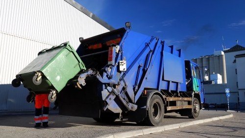 A large blue refuse collection truck with hydraulic lifting arms and an enclosed cab is in the process of emptying a green wheeled waste bin into its rear hopper. The green bin appears to be made of durable plastic, with a textured surface and fitted with black wheels and a lid. The truck is parked on a paved surface in an urban setting, with a white industrial building featuring vertical siding on the left and additional modern white and grey structures in the background, under a clear blue sky. A worker in high-visibility orange trousers and a dark jacket is partially visible near the waste bin, assisting with the rubbish collection. The scene is well-lit by natural daylight, emphasizing the clean, crisp coloring of the truck and surrounding environment, typical of rubbish removal services offered in the Notting Hill area, as managed by Notting Hill House Clearance.