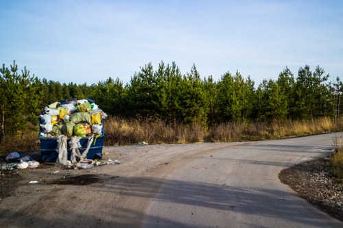 A large blue waste skip situated on the side of a rural dirt road, filled with various black, yellow, and clear plastic bags containing rubbish, along with loose debris and discarded materials spilling onto the ground. The skip appears to be used for rubbish removal services, with items such as packaging and general household waste visible. In the background, there is a line of green pine trees under a pale blue sky, indicating a natural outdoor setting. The dirt road curves gently to the right, with patches of dried grass and gravel surrounding the skip, showing signs of recent use. This scene relates to rubbish collection or clearance services provided in the Notting Hill area, highlighting the importance of professional waste removal for proper disposal of household or garden waste.