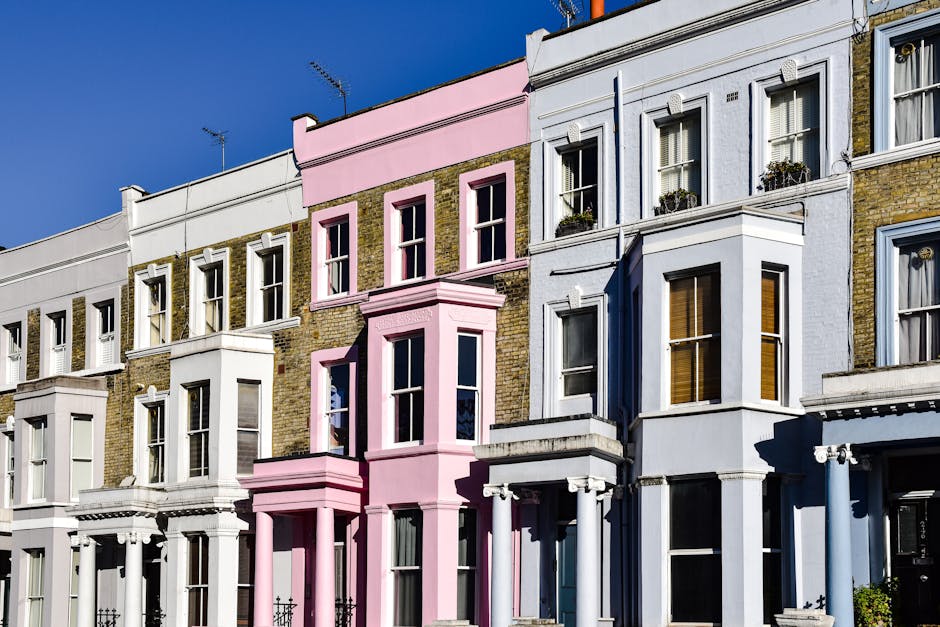 A row of multi-story Victorian terraced houses with a uniform façade, each painted in pastel shades of white, pink, red, and light blue. The buildings feature intricate architectural detailing such a
